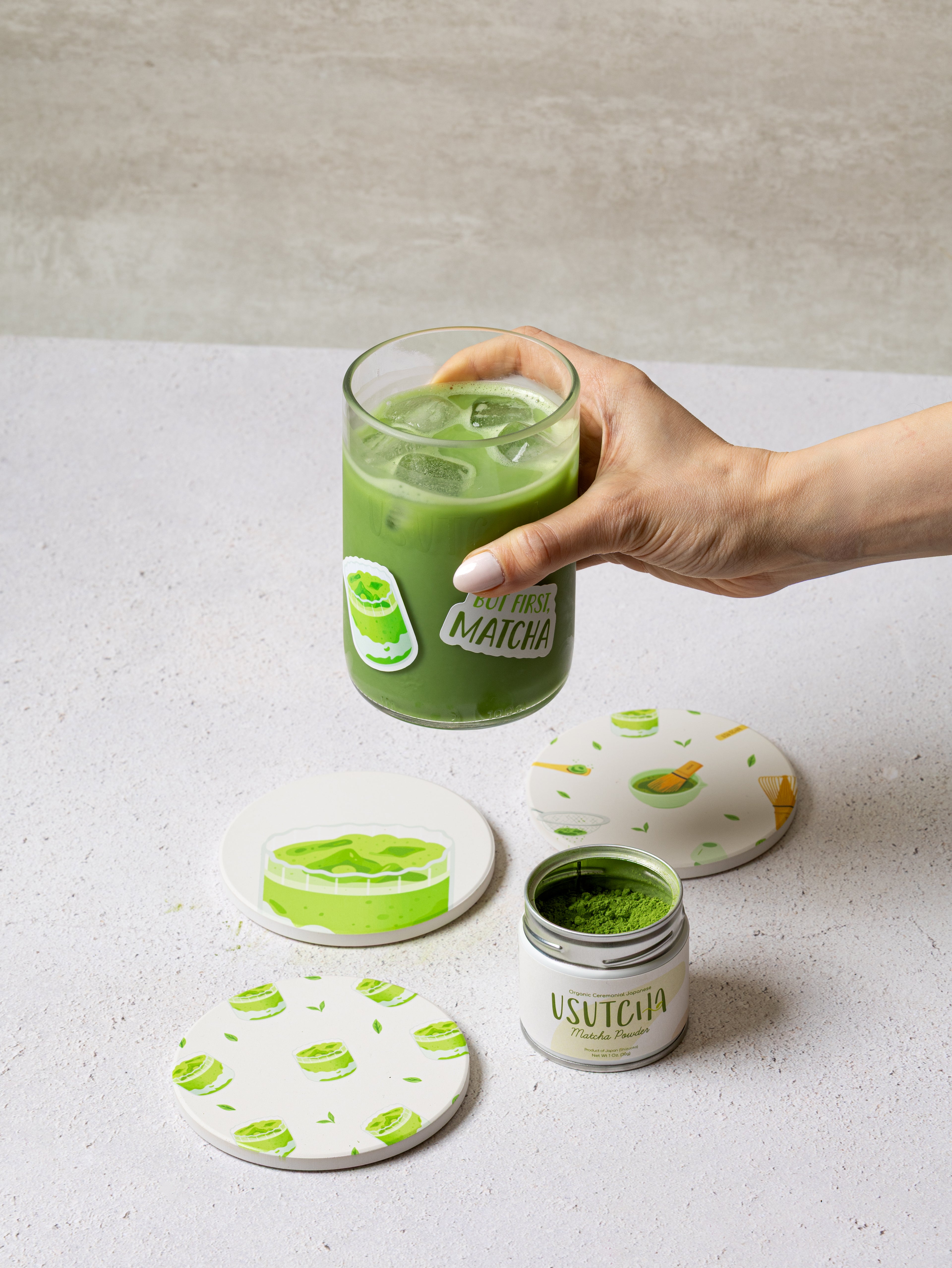 Hand holding a glass of iced green matcha drink with a container of Usucha matcha powder on a light surface.